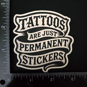 May include: A black and white sticker featuring the text "TATTOOS ARE JUST PERMANENT STICKERS" in a stylized font on a ribbon design. A ruler is visible along the left and bottom edges, showing the sticker is approximately 2.5 inches wide.