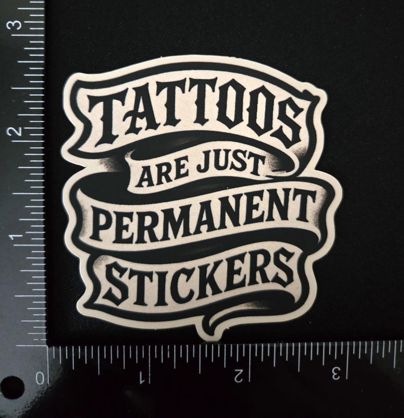 May include: A black and white sticker featuring the text "TATTOOS ARE JUST PERMANENT STICKERS" in a stylized font on a ribbon design. A ruler is visible along the left and bottom edges, showing the sticker is approximately 2.5 inches wide.