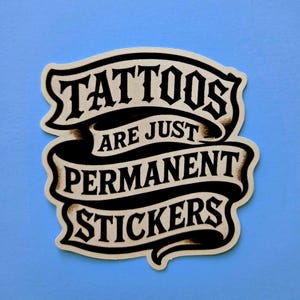 May include: A die-cut sticker featuring a black and cream-colored banner design with the text TATTOOS ARE JUST PERMANENT STICKERS in a bold, stylized serif font. The graphic has a vintage-inspired aesthetic with dark shading and curved edges.