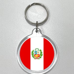 Puede incluir: Un llavero redondo con la bandera de Perú. El diseño de la bandera incluye franjas verticales rojas y blancas con el escudo de armas peruano en el centro. El llavero tiene un anillo de metal plateado.