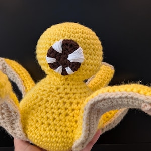 Può includere: Un polpo giallo all'uncinetto con un occhio marrone e bianco. Il polpo ha sei tentacoli con un bordo marrone.