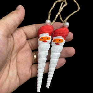 Könnte beinhalten: Zwei Santa Claus-Ornamente aus weißen Spiralmuscheln, jeweils mit roter Mütze, orangefarbenem Gesicht und rotem Schnurrbart. Sie hängen an einer natürlichen Schnur mit einer Perlenperle. Die Ornamente sind vor schwarzem Hintergrund.
