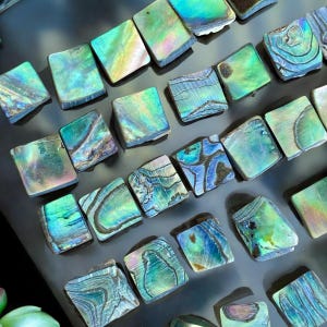 Könnte beinhalten: Nahaufnahme von quadratischen Abalone-Muschelteilen. Die Muscheln zeigen schillernde Farben von Grün, Blau und Lila. Jedes Stück hat ein einzigartiges, natürliches Muster und Textur. Die Muscheln sind mosaikartig angeordnet.