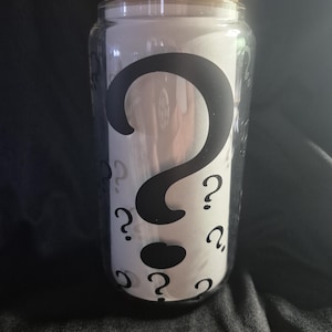 Könnte beinhalten: Ein klares Glas mit einem Bambusdeckel, verziert mit einem großen schwarzen Fragezeichen und kleineren Fragezeichen. Das Glas ist mit einem weißen Material gefüllt.