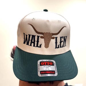 May include: A beige and green baseball cap with a brown bull's head and horns embroidered on the front. The cap has the text "WAL LEN" embroidered on the front. The cap has a label on the back that says "OTTO" in red letters. The label also says "CLASSIC FIT OSFM" and "MID PROFILE MAXIMUM COMFORT".