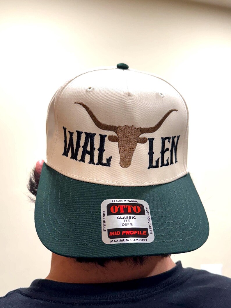 May include: A beige and green baseball cap with a brown bull's head and horns embroidered on the front. The cap has the text "WAL LEN" embroidered on the front. The cap has a label on the back that says "OTTO" in red letters. The label also says "CLASSIC FIT OSFM" and "MID PROFILE MAXIMUM COMFORT".