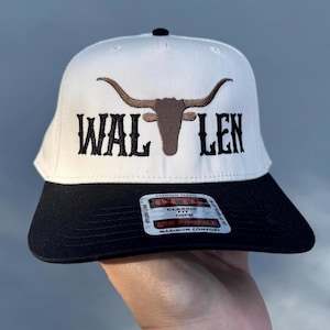 May include: White baseball cap with a black brim. The front of the cap features a brown embroidered bull's head with horns and the text "WAL LEN" in black embroidery.
