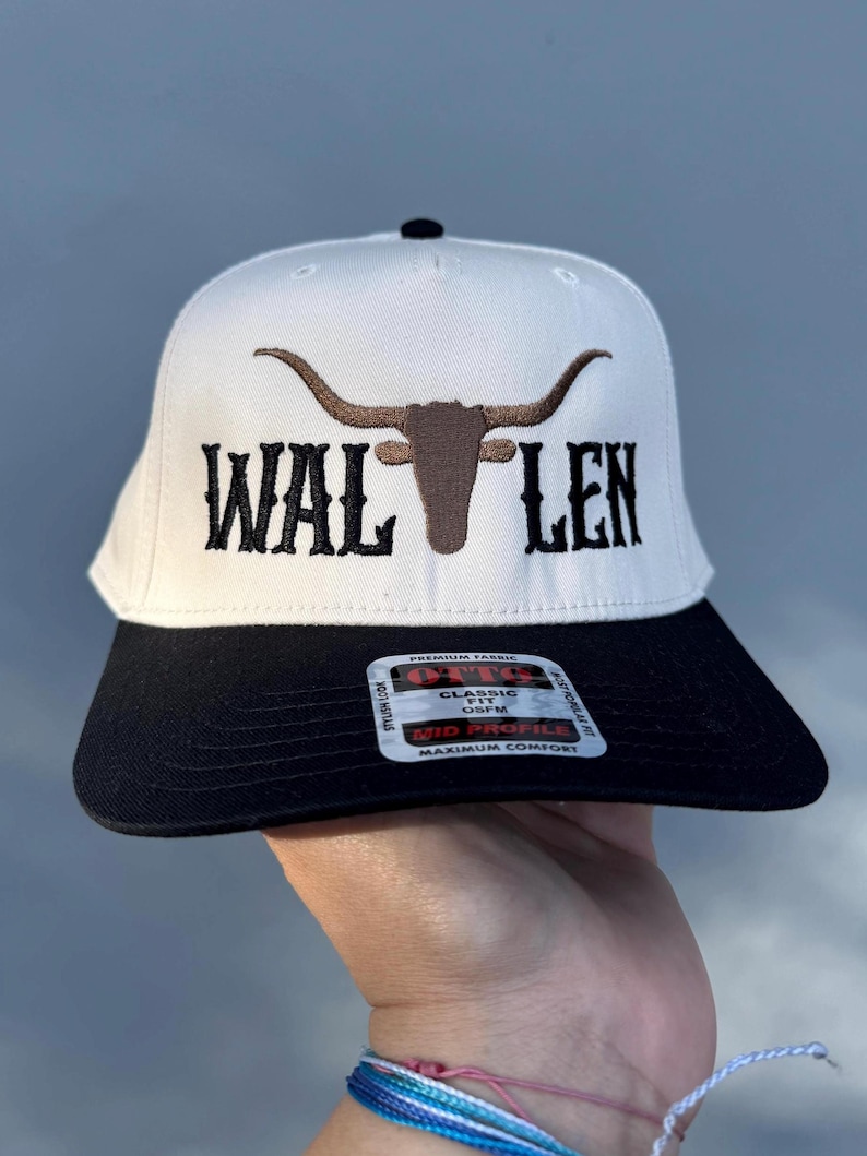 May include: White baseball cap with a black brim. The front of the cap features a brown embroidered bull's head with horns and the text "WAL LEN" in black embroidery.