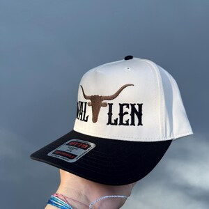 May include: White and black baseball cap with a brown bull's head and the text "VAL LEN" embroidered on the front.