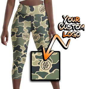 May include: Camouflage print capri leggings with a high waist and a customizable logo area. The logo area is highlighted with an orange arrow. The leggings are a light brown and green color.