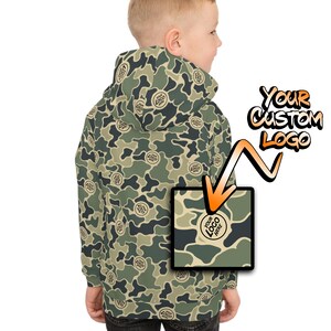 May include: A camouflage-patterned hoodie in shades of green, brown, and tan. The hoodie features a repeating pattern of abstract shapes and a hood. The text "YOUR CUSTOM LOGO" is displayed in a stylized font. A small square shows a sample logo.