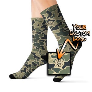 May include: A pair of crew socks with a camouflage pattern in shades of green, brown, and black. The socks have black heels and toes. The text "YOUR CUSTOM LOGO" is displayed on the image.