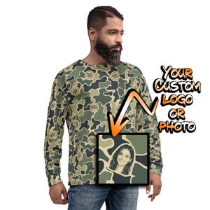 May include: A long-sleeve sweatshirt featuring an all-over green, tan, and black camouflage pattern with a repeating circular logo design. An orange arrow points to a square inset displaying a portrait of a smiling person. Text reads YOUR CUSTOM LOGO OR PHOTO.