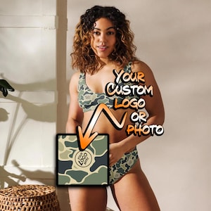 May include: A two-piece bikini set featuring a green, tan, and dark gray camouflage pattern. The top is a scoop-neck sports bra style, and the bottom is a high-cut brief. An overlay graphic shows the text YOUR CUSTOM LOGO OR PHOTO.