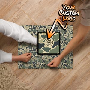 May include: Two people are putting together a camouflage jigsaw puzzle. The puzzle has a space for a custom logo. The text "YOUR CUSTOM LOGO" is written in a speech bubble above the puzzle.