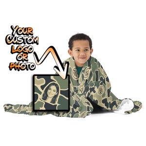 May include: A camouflage patterned blanket featuring a repeating print of a circular logo with the text YOUR LOGO HERE and a square inset displaying a stylized portrait of a person, with the text YOUR CUSTOM LOGO OR PHOTO and an arrow.