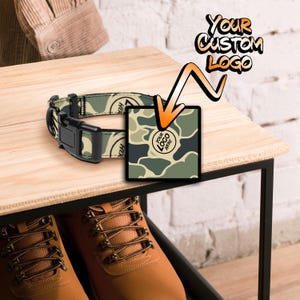 May include: A camouflage dog collar with a black buckle and a space for a custom logo. The collar is on a wooden surface with a pair of brown leather shoes in the background.