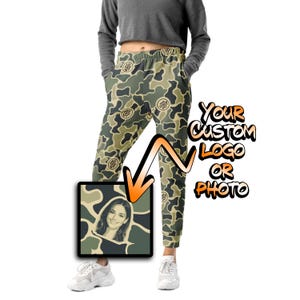 May include: Camo print joggers featuring a repeating circular logo pattern in shades of olive green, tan, and dark brown. An inset square graphic displays a portrait of a smiling person against a camo background with an orange arrow pointing to it.