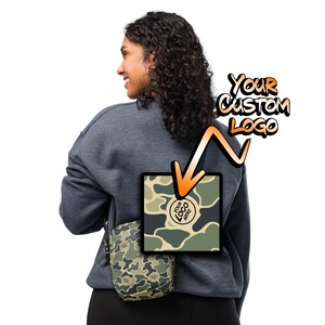 May include: A person wearing a gray sweatshirt with a camouflage crossbody bag. The bag has a circle on the front that says "YOUR LOGO HERE". The text "YOUR CUSTOM LOGO" is written in orange above the bag.