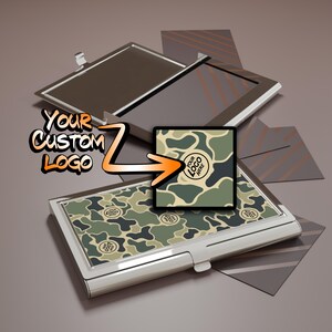 May include: A silver business card holder with a camouflage pattern. The holder is open, revealing a mirrored interior. The text "YOUR CUSTOM LOGO" is displayed with an arrow pointing to the camouflage design.