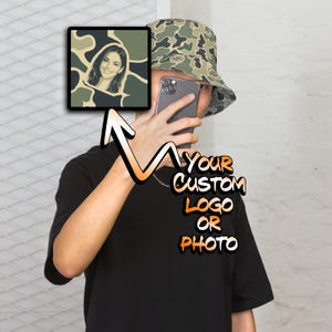 May include: A camouflage bucket hat with a dark gray smartphone held in front of the face. A square graphic shows a portrait of a person against a camo background. The text "YOUR CUSTOM LOGO OR PHOTO" is displayed in orange and white lettering.