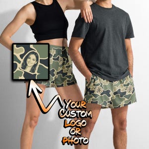 May include: A pair of camouflage print shorts featuring a repeating pattern of olive green, dark green, and tan shapes. The shorts include a circular logo graphic on the fabric. The text "YOUR CUSTOM LOGO OR PHOTO" is displayed in the image.