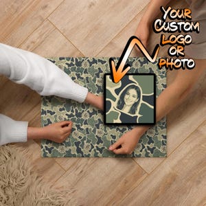 May include: A rectangular jigsaw puzzle featuring a camouflage pattern in shades of green, tan, and dark gray. A square section in the center displays a portrait of a person in a stylized graphic format. The text "YOUR CUSTOM LOGO OR PHOTO" is visible.