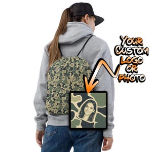 May include: A drawstring backpack featuring a green, brown, and tan camouflage pattern with black cord straps. A square inset displays a portrait of a smiling person against a camo background. Text reads: Your Custom Logo Or Photo.