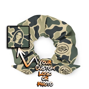 May include: A fabric hair scrunchie featuring a camouflage pattern in shades of olive green, dark grey, and tan. The scrunchie includes a square inset displaying a portrait of a person and circular areas with the text YOUR LOGO HERE.