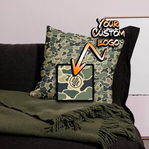 May include: A green camouflage pillow with a white circle that says "YOUR LOGO HERE" in the center. The pillow is on a couch with a green blanket.