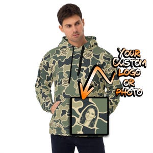 May include: A camouflage print hoodie featuring a repeating logo pattern and a large square graphic on the front pocket area. The graphic displays a portrait of a person and an orange arrow pointing toward it, with text reading YOUR CUSTOM LOGO OR PHOTO.