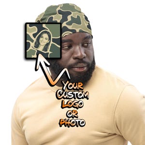 May include: A camouflage beanie featuring a square graphic insert showing a portrait of a smiling person. Below the graphic, the text reads "YOUR CUSTOM LOGO OR PHOTO" in a stylized orange and black font, indicating a customizable product option.
