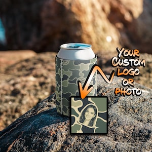 May include: A camouflage print foam can cooler sleeve featuring a white top rim and a vertical white stitched seam. An orange arrow points to a square graphic insert displaying a portrait of a person with the text YOUR CUSTOM LOGO OR PHOTO.