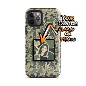 May include: A smartphone case featuring a green, brown, and tan camouflage pattern with small circular 'YOUR LOGO HERE' text. A square attachment in the center displays a portrait of a smiling person. An orange arrow points to the square.