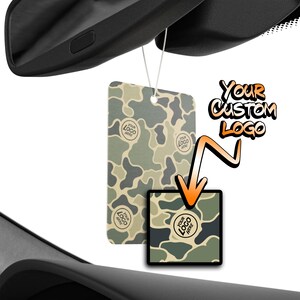 May include: A rectangular car air freshener hanging from a rearview mirror, featuring a green, tan, and dark gray camouflage pattern with circular 'Your Logo Here' text. An orange arrow points to a square version of the same camouflage design.