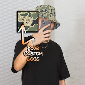 May include: A person wearing a black t-shirt with the text "YOUR CUSTOM LOGO" and a camouflage bucket hat. The hat has a white circle with the text "YOUR LOGO HERE" in black.