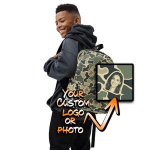 May include: A camouflage print backpack featuring a repeating circular logo pattern. The backpack includes a square graphic insert displaying a portrait of a smiling person against a camo background. Text on the image reads Your Custom Logo Or Photo.