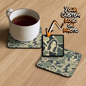 May include: Two square coasters with a green, tan, and dark gray camouflage pattern featuring the text "YOUR LOGO HERE". One coaster supports a white ceramic mug filled with dark liquid, while the other displays a custom portrait graphic.
