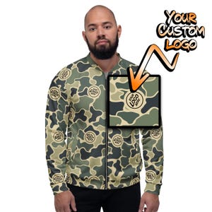 May include: A green, brown, and beige camouflage bomber jacket with a zipper closure. The jacket has a black circle with the text "YOUR LOGO HERE" repeated throughout the pattern. The text "YOUR CUSTOM LOGO" is written in orange and black on a white background.
