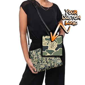 May include: A green and brown camouflage print crossbody bag with a black strap. The bag has a space for a logo that says "Your Logo Here".