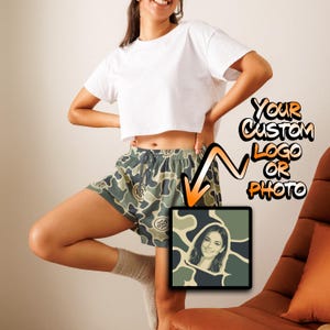 May include: A pair of green and dark gray camouflage print shorts with an elastic drawstring waistband. The shorts feature a customizable area for a logo or photo, shown with an example graphic of a person's portrait within the camo pattern.