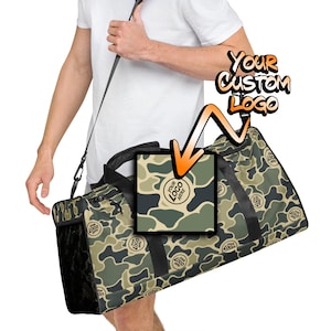 May include: A camouflage print duffel bag with a black strap and black accents. The bag has a space for a custom logo. "Your Custom Logo" is written in a graffiti style font.