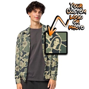 May include: A camouflage print zip-up jacket featuring a repeating circular logo pattern and a square graphic insert displaying a portrait of a person. The text "YOUR CUSTOM LOGO OR PHOTO" appears above the graphic with an orange arrow pointing down.