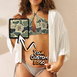 May include: A two-piece swimsuit with a camouflage pattern. The top has a halter style with a cutout in the front. The bottom is a simple bikini style. The swimsuit is shown on a model with a white shirt over it. The text "YOUR CUSTOM LOGO" is written on the model's stomach.