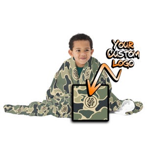 May include: A child wearing a green and brown camouflage print blanket with a white hood. The blanket has a space for a custom logo. The text "Your Custom Logo" is written in orange and black.
