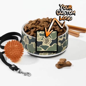 May include: A stainless steel dog bowl with a camouflage pattern and a space for a custom logo. The bowl is filled with dog food and there is a dog leash, a dog toy, and dog treats in the background.