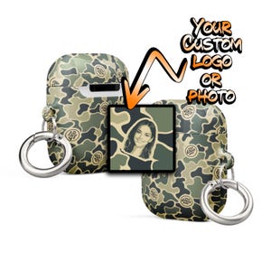 May include: Two camouflage-patterned protective cases for wireless earbuds, each featuring a silver metal ring keychain. A central square graphic displays a portrait of a person, with text above reading YOUR CUSTOM LOGO OR PHOTO.