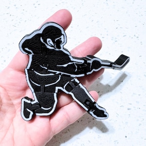 May include: Black and white hockey player figurine in a crouching position, holding a hockey stick. The figurine has a textured surface and a white outline. The hockey player is in a dynamic pose, suggesting action.