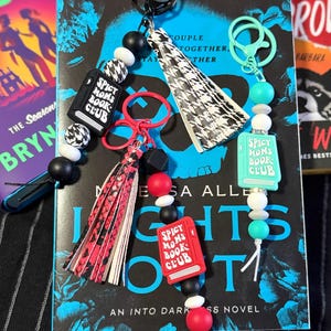 May include: Keychains with book-themed charms and beads. The keychains feature charms that say "Spicy Moms Book Club" and a tassel. The background includes a book cover with the title "Lights Out" and the text "#1 New York Times Bestseller".