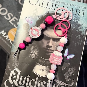 May include: Pink and white beaded keychain with charms that read "Bad Bitch Book Club" and "Crying Over Fictional Characters." The keychain is displayed over a book titled "Quicksilver" by Callie Hart.